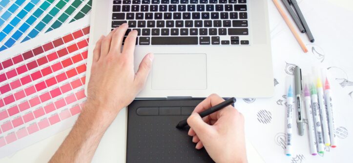 Graphic designer working on a laptop with a trackpad, colour charts and markers beside them