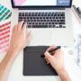Graphic designer working on a laptop with a trackpad, colour charts and markers beside them